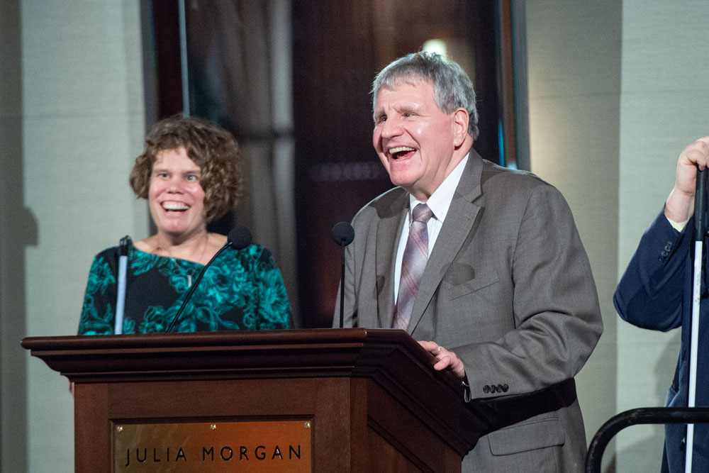 George Kerscher at podium for acceptance with Erin Lauridsen, Director, Access Technology from the Lighthouse standing in the background. Erin introduced the award.