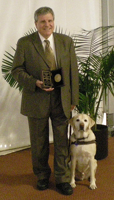 George Kerscher holding the Migel Medal award with Mikey his guide dog by his side.